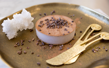 Load image into Gallery viewer, A dessert plate featuring a round chocolate mousse dusted with cocoa and sprinkled with cacao nibs, accompanied by a gold dessert fork and a cluster of white sugar crystals.

