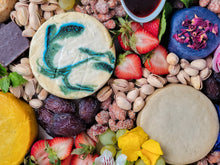 Load image into Gallery viewer, close up of an overflowing cheese board with nuts, dates, berries, flowers and cheese wheels

