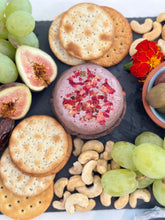 Load image into Gallery viewer, A wheel of vegan cheese topped with edible flowers, surrounded by fresh figs, grapes, crackers, and nuts on a slate board.
