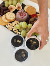 Load image into Gallery viewer, A hand placing two containers of SriMu vegan cheese near a platter with fruits, nuts, and crackers.
