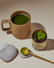 Load image into Gallery viewer, A cup of matcha tea next to an open jar of matcha powder and a golden spoon with powder.
