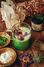 Load image into Gallery viewer, A glass of matcha latte garnished with purple flowers, next to a jar of matcha powder and a bamboo whisk on a wooden surface.
