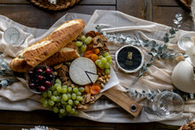 Load image into Gallery viewer, Cheese board seen from above with lots of fruit, two cheese wheels and baguette
